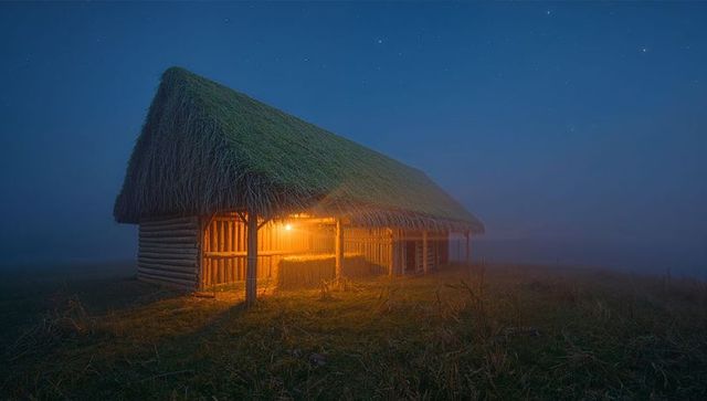 Glowing thatch-roof barn standing in misty meadow at night revealing hay bales