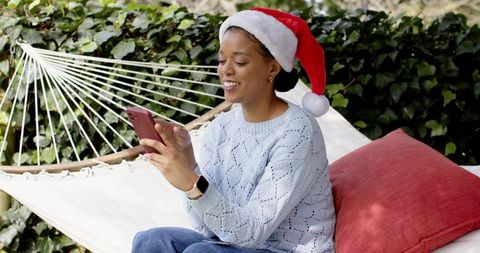 African american woman relaxing in hammock wearing santa hat while using smartphone
