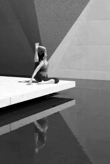 Woman wearing sports attire practicing yoga near calm water using a concrete platform for stability. Ideal for websites, promotional materials or wellness blogs focusing on fitness, meditation, and mental well-being. Monochrome tones enhance sense of tranquility and focus, perfect for calmness or minimalism themes.