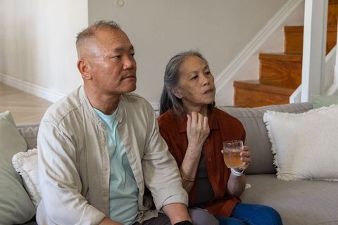 Asian senior couple sitting on sofa holding drink, cozy home companionship and relaxation