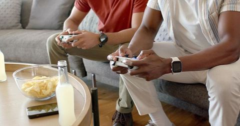 Two friends gaming on sofa with controllers, snacks and drinks in cozy home lounge