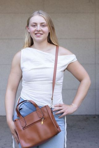 Young woman smiling wearing white one-shoulder top holding brown leather crossbody bag