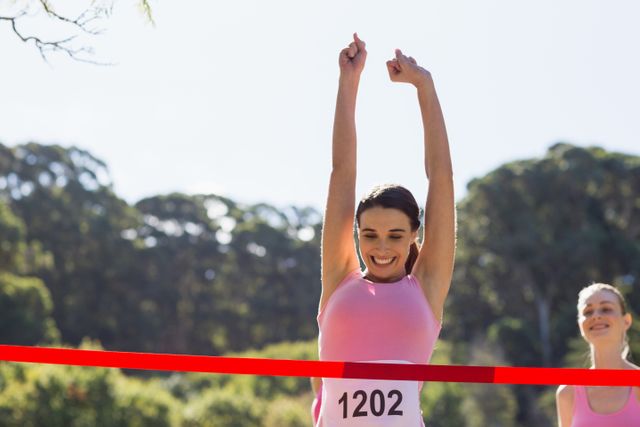 This image depicts a joyful female athlete triumphantly crossing the finish line with her arms raised in celebration during an outdoor race in a park. Ideal for use in articles or promotions related to sports, fitness, motivation, athletic competitions, and active lifestyle campaigns. The bright, sunny setting and the athlete's visible joy highlight themes of success and determination.