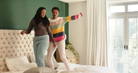 Women Joyfully Bouncing on Bed by Sunlit Window in Cozy Bedroom