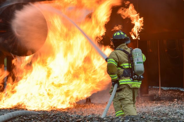 Firefighter dressed in full protective gear courageously fighting a fierce fire using a water hose. Suitable for use in topics related to emergency services, firefighting, safety measures, and first responder training. Can be used in articles or presentations on fire safety, public service announcements, and educational material for emergency response preparedness.