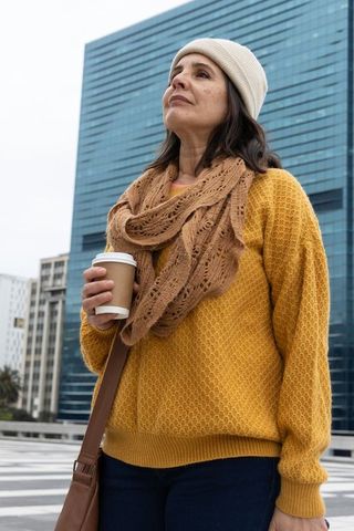 Urban woman standing with coffee and knit scarf by blue-glass tower, gazing upward