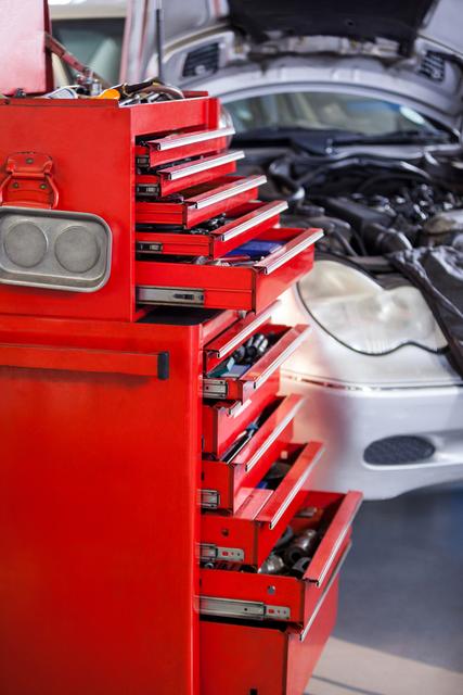 Red toolbox with open drawers filled with various tools in an auto repair garage. Car in background with open hood. Ideal for illustrating automotive repair services, mechanic workshops, and maintenance facilities.