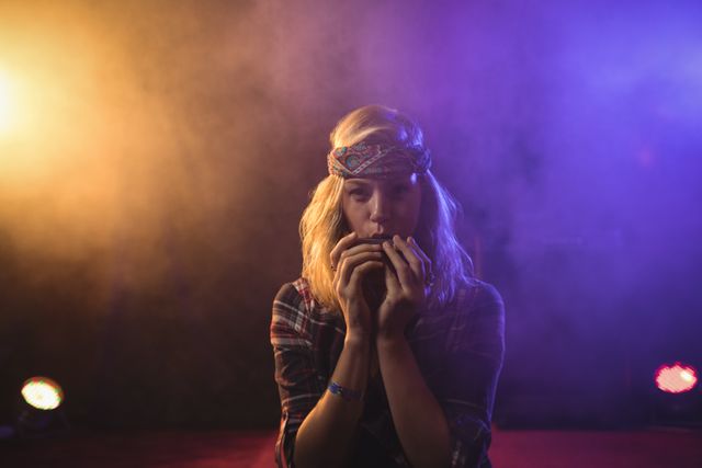 Female musician playing harmonica on stage with colorful lights and smoke effects. Ideal for use in articles about live music, nightlife, entertainment, and artistic performances. Can be used for promotional materials for music events, concerts, and nightclubs.