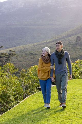 Asian couple walking hand-in-hand on hillside lawn wearing scarves and knitwear