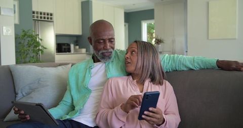 Senior couple relaxing on couch sharing tablet and smartphone in modern open-plan home
