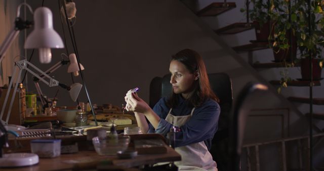 A woman is crafting jewelry in her home workshop at night. She is deeply focused, examining an intricate piece. The lighting and décor suggest a cozy, intimate ambiance perfect for creative work. This image is ideal for use in articles or advertisements about small businesses, crafts, creative pursuits, or work-from-home environments.