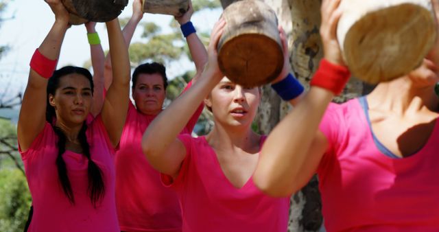 Group of women participating in an outdoor fitness boot camp lifting logs above their heads. They are wearing pink shirts and wristbands, showing determination and teamwork. Perfect for articles on fitness, outdoor activities, and group workouts. Suitable for promoting fitness communities, boot camp classes, or healthy lifestyle campaigns.