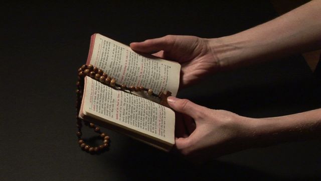 A pair of hands holding an open prayer book intertwined with rosary beads against a dark background. Ideal for illustrating themes of faith, spirituality, devotion, prayer, and religious contemplation. This visual can be used in religious studies materials, spiritual guides, blogs about faith, and worship-related publications.