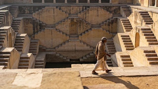 Elderly man walking along sunlit historic stepwell ledge with geometric stone terraces