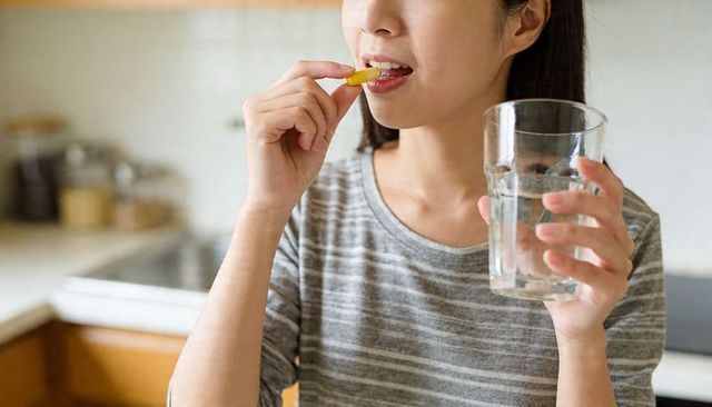 Young woman taking yellow supplement capsule while holding glass of water in home kitchen