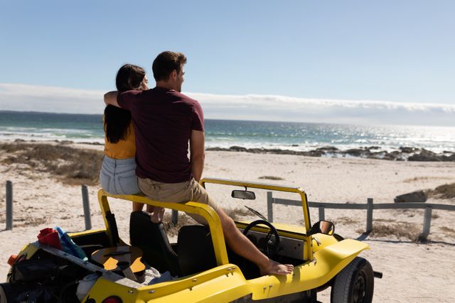 Couple sitting on yellow beach buggy, embracing and admiring ocean view. Ideal for travel blogs, romantic getaway promotions, summer vacation advertisements, and lifestyle magazines.