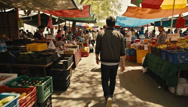 Man walking through bustling outdoor market under colorful tarps with fruit stalls