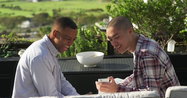 Two men sit on an outdoor sofa, enjoying each other's company. One holds a smartphone, likely sharing something amusing, while both are smiling and engaged in conversation. This image captures the essence of friendly bonding and relaxation, making it perfect for themes around friendship, casual get-togethers, social interactions, and outdoor leisure.