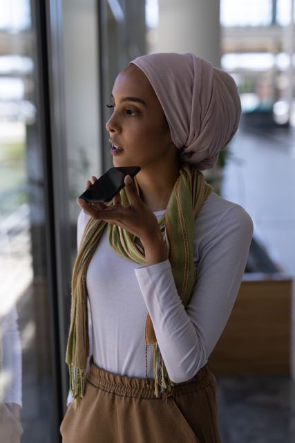 Biracial woman with light brown skin and dark eyes speaking into a smartphone in an office environment. She is wearing a pink headscarf, a white top, and a scarf around her neck, giving a casual yet professional look. This image can be used for business communication, technology, modern workspaces, and diversity in the workplace themes.