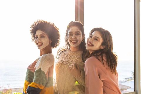 Diverse women wearing clay face masks enjoying coastal sunlit self-care moment by window
