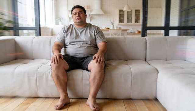 Man sitting on beige sectional sofa wearing gray tee and shorts in sunlit open-plan living room