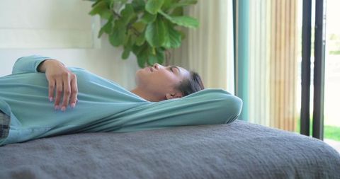 Asian woman resting on bed in teal top enjoying tranquil daylight and indoor greenery