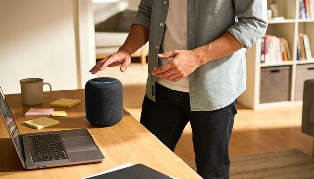 Casual man reaching and tapping smart speaker on home desk with laptop and coffee
