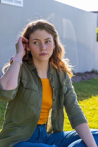 Young woman sitting on grass touching hair in sunlit casual olive jacket and denim with smartwatch