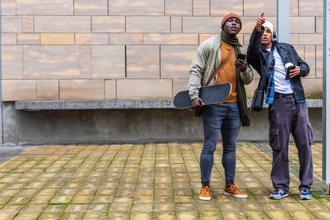 Urban friends pointing while holding skateboard and coffee, casual youth lifestyle scene
