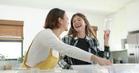 Laughing friends organizing clear pantry jars and containers in bright modern kitchen
