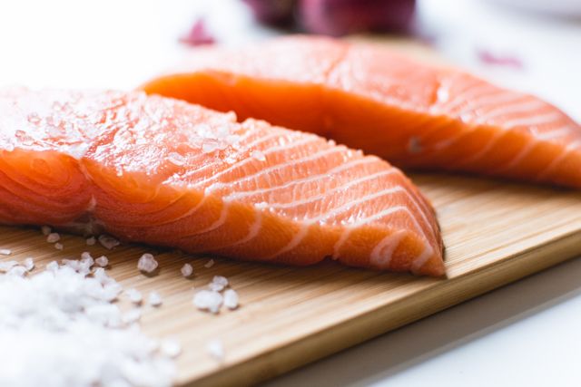 High-quality stock photo depicting fresh raw salmon fillet slices placed on a wooden cutting board with a scattering of sea salt crystals. Ideal for use in health, nutrition, cooking, or food preparation content. Suitable for illustrating seafood recipes, restaurant menus, blog posts about healthy eating, and culinary magazines.