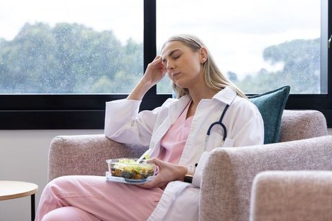 Tired female doctor taking break with salad rubbing temple nurse burnout concept
