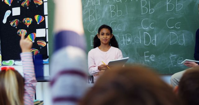 Young girl presenting in front of a classroom with a chalkboard and letters in the background. Classmates eagerly raising hands to participate in discussion. Ideal for content about education, classroom learning, student engagement, teaching methods, school activities.