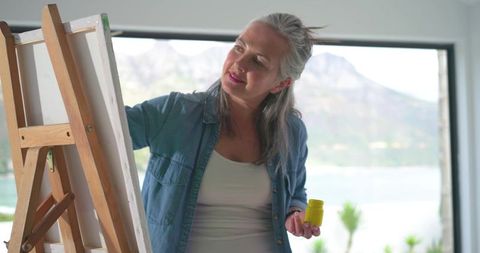 Mature woman painting on canvas in bright lakeside studio, holding yellow paint jar