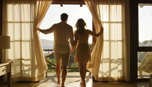 Couple holding hands pulling curtains to seaside balcony at golden hour backlit silhouette