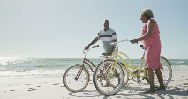 Senior couple walking with bikes on the sandy shoreline during a sunny day by the ocean. Ideal for themes related to retirement, active lifestyle, beach vacations, carefree living, and exercise.