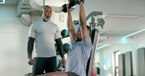 African american trainer spotting client lifting dumbbells on incline bench in modern gym