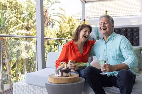 Senior couple celebrating 50th birthday on sunny balcony laughing with cake and wine