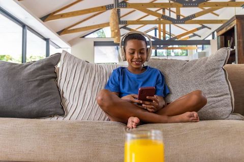African american boy sitting on sofa using smartphone and headphones in bright living room