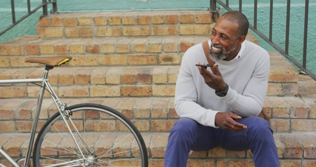 Older man in casual clothes resting on brick steps outdoors using smartphone with smile, bicycle next to him. Ideal for themes like active lifestyle, senior digital engagement, urban living, relaxation.