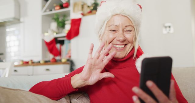 Happy senior caucasian woman wearing santa claus hat, using smartphone for image call. Spending quality time at home alone at christmas.