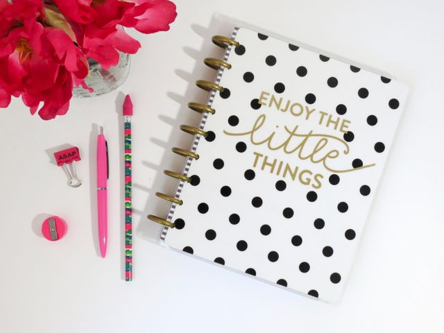 Polka dot notebook with 'Enjoy the Little Things' message in gold lettering accompanied by pink stationery, including a pen, pencil, eraser, and a binder clip, placed on a white desk. Perfect for illustrating concepts of goal setting, planning, creative workspace design, organization, or inspirational stationery. Ideal for use in office supply advertisements, productivity blogs, or academic planning resources.