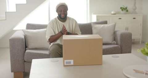 Joyful african man unboxing delivery box on living room table, modern minimalist interior