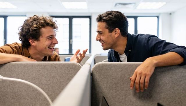 Two coworkers leaning over cubicle dividers smiling and chatting in modern open office