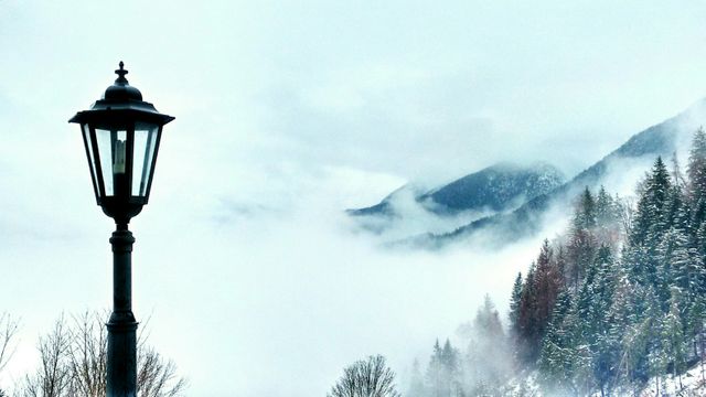 Snow-covered mountain scenery featuring a solitary street lamp. Fog drifts among snowy trees, creating a serene and peaceful atmosphere. Can be used in marketing materials for travel destinations, winter tourism promotions, vacation planning resources, or inspirational nature blog posts.