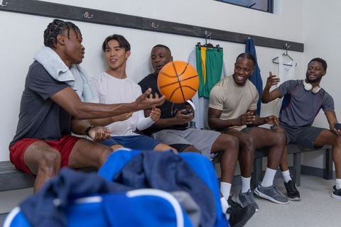 Diverse male basketball teammates laughing and passing basketball in locker room camaraderie