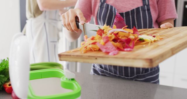 This image depicts a person wearing an apron composting kitchen scraps into a compost bin. Colorful vegetable peels and food waste are being scraped off a wooden cutting board. Ideal for use in articles or blogs promoting eco-friendly living, sustainable household practices, food preparation, and zero waste lifestyles. Useful for campaigns related to environmental awareness and healthy home environments.