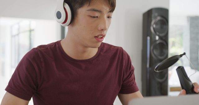 Young man wearing headphones, sitting at home, recording a podcast using his microphone. Ideal for content centered on podcasting, home studios, young creators, audio production, and modern technology use in everyday life.