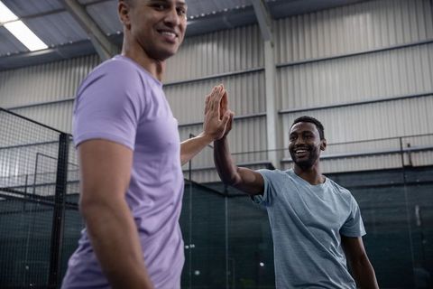 High-fiving male friends celebrating teamwork on indoor court in sports hall under skylight