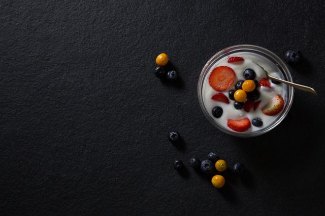 Top view of a bowl filled with yogurt, strawberries, blueberries, and other fresh berries on a black background. Ideal for use in articles or advertisements about healthy eating, nutritious breakfast options, wellness, and diet plans. Perfect for food blogs, recipe websites, and health-focused publications.